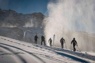 Skibergsteiger im Aufstieg über eine Kunstschneepiste vor einer Bergkulisse.