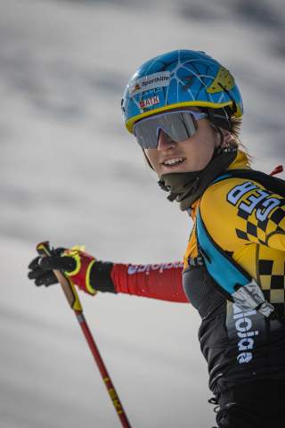 Porträt einer Skifahrerin mit blauem Helm, Skibrille und gelber Sportbekleidung, die einen Skistock hält und in verschneiter Umgebung unterwegs ist.