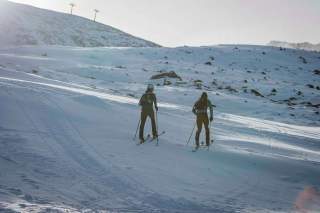 Zwei Skifahrer mit Helmen und Rucksäcken laufen mit Skistöcken einen schneebedeckten Hang hinauf, umgeben von einer verschneiten Berglandschaft.