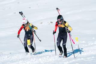 Zwei Skibergsteiger laufen mit Ski am Rucksack eine Piste hinauf.
