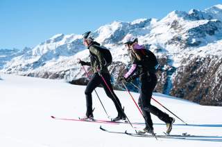 Zwei Skitourengeher steigen mit Skiern und Stöcken einen verschneiten Hang hinauf, im Hintergrund schneebedeckte Berge und blauer Himmel.