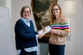 Zwei lächelnde Frauen mit Brillen übergeben sich ein Buch mit dem Titel „Darum Berge“. Beide tragen Pullover und stehen vor einem unscharfen Hintergrund mit Text.