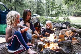 Drei Kinder sitzen am Lagerfeuer und warten auf das Essen auf dem Grill.