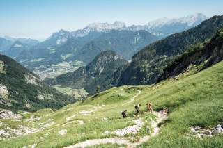 Drei Wanderer steigen einen schmalen Pfad auf einer grünen Bergwiese hinauf, im Hintergrund weite Aussicht auf bewaldete Berge und ein Tal.