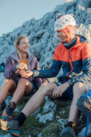 Zwei Bergsteiger, eine Frau und ein Mann mit Kletterhelm, sitzen auf felsigem Gelände und lachen, während sie sich etwas zu essen reichen.