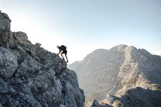 Eine Person mit Rucksack und Helm klettert über einen felsigen Grat; dahinter steile, helle Berggipfel unter blauem Himmel.