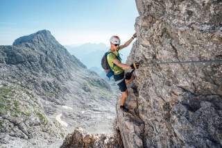 Ein Bergsteiger mit Helm und Kletterausrüstung erklimmt gesichert eine steile Felswand in alpiner Berglandschaft bei klarem Himmel.