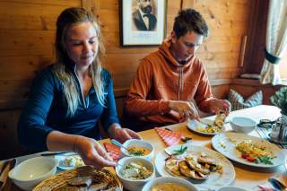 Zwei Personen sitzen an einem Holztisch in einem rustikalen Raum und essen gemeinsam verschiedene Gerichte wie Brot, Aufstriche und Pfannkuchen.