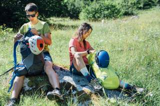 Zwei Personen sitzen auf einem Stein in einer Wiese und packen ihre Rucksäcke aus, an denen Helme befestigt sind.