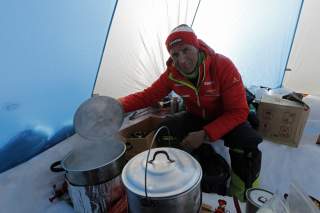 Ralf Dujmovits sitzt im Zelt beim Kochen mit zwei großen Töpfen. Er trägt Winterklamotten, um ihn herum liegt Schnee.