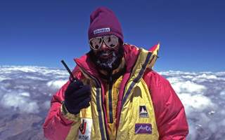 Ralf Dujmovtis steht in Expeditionsausrüstung auf dem K2 und hält ein Walkie Talkie in der Hand.