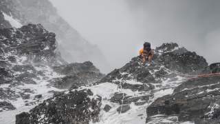 Ein Mensch in Expiditionsausrüstung klettert über verschneite Felsen bei Nebel und Schneefall.