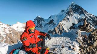 Tamara Lunger in Expeditionsmontur pausiert bei strahlendem Sonnenschein im Schnee auf Felsen.