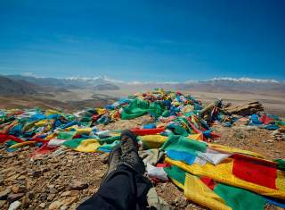 Verschränkte Beine mit Bergschuhen an den Füßen vor Hochgebirgskulisse im Himalaya und vielen Gebetsfahnen am Boden