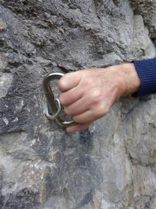 Eine Hand hält einen Karabiner, der in einen geklebten Bohrhaken eingehängt ist, um durch Drehversuch zu Testen, ob der Haken sich bewegt und damit nicht sicher ist.