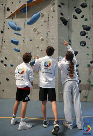 Drei Jugendliche in Sportkleidung stehen vor einer Kletterwand in der Halle und zeigen auf verschiedene Griffe.