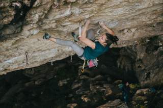 Frau klettert an einer steilen Felswand, gesichert mit Kletterausrüstung und Chalkbag, im Hintergrund Felsen und eine weitere Person.