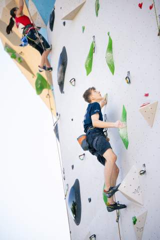 Zwei Personen klettern an einer künstlichen Kletterwand, eine junge Frau oben links und ein junger Mann unten rechts, beide gesichert mit Klettergurten.