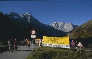 Menschen stehen auf einem Wanderweg in den Bergen und halten ein gelbes Banner mit der Aufschrift „Mountain Wilderness – Für eine autofreie Eng“, daneben Verkehrsschilder, im Hintergrund schneebedeckte Gipfel.