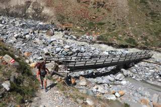 Wanderin vor einer zerstörten Holzbrücke über einen steinigen Wildbach, im Hintergrund weitere Wanderer und eine intakte Brücke.