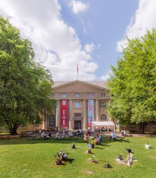 Historisches Gebäude mit Säulen und roter Fahne, davor eine Wiese mit Bänken, Menschen und Blumenarrangements, umgeben von Bäumen.