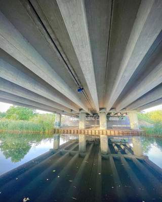 Blick von unten auf eine Betonbrücke über einem ruhigen Gewässer, das die Brücke und die umliegende Natur spiegelt.