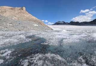 Gletscherlandschaft mit schmelzendem Eis und Wasser im Vordergrund, felsigem Hang links und einer Berghütte auf dem Hügel, Berggipfel und blauer Himmel im Hintergrund.