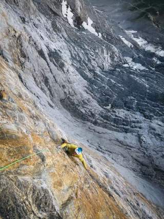 Bergsteiger mit gelber Jacke und blauem Helm klettert an einer steilen Felswand, mit Seil gesichert.