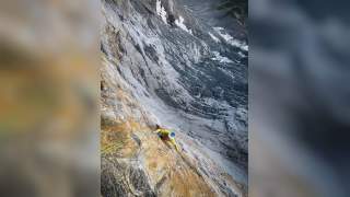 Bergsteiger klettert an einer steilen Felswand, im Hintergrund strahlende Sonne und schneebedeckter Gipfel unter blauem Himmel.
