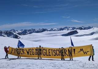 Mehrere Personen stehen auf einem verschneiten Gletscher und halten ein großes gelbes Banner mit der Aufschrift „Finger weg von den Gletschern!“, im Hintergrund schneebedeckte Berge und blauer Himmel.