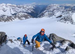 Drei Bergsteiger in Winterkleidung und mit Eispickeln klettern einen verschneiten Berghang hinauf, im Hintergrund schneebedeckte Alpen.