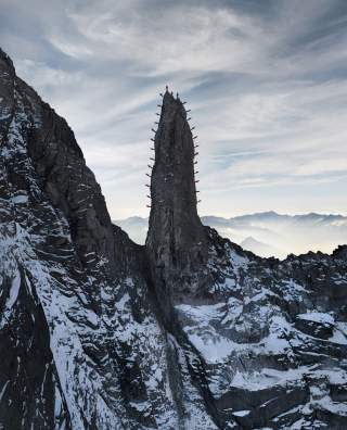 Spitzer, steiler Felsen in einer winterlichen Gebirgslandschaft, auf dessen Rand mehrere Menschen in roten Jacken stehen.