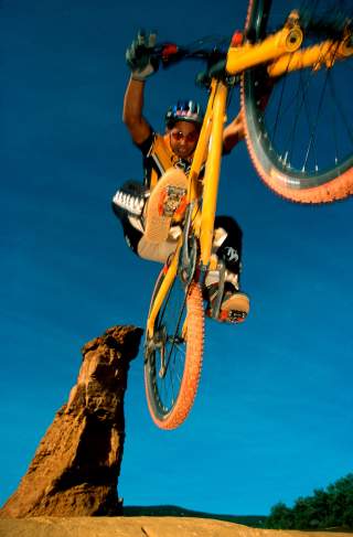 Mountainbiker mit gelbem Fahrrad springt über Felsen vor blauem Himmel.