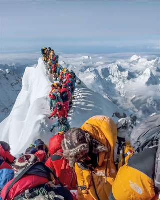 Eine lange Reihe von Bergsteigern in bunter Ausrüstung steht auf einem schmalen, verschneiten Grat in großer Höhe, umgeben von schneebedeckten Bergen.