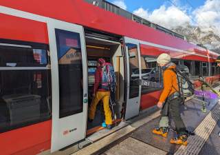Zwei Menschen in Skikleidung und mit Skiausrüstung steigen in einen roten Regionalzug der Deutschen Bahn ein.