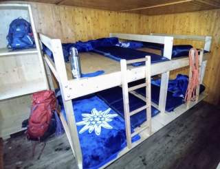 Ein großes Doppelstockbett aus Holz auf einer Hütte. Blaue Decken mit einem weißen Edelweiß liegen darauf.