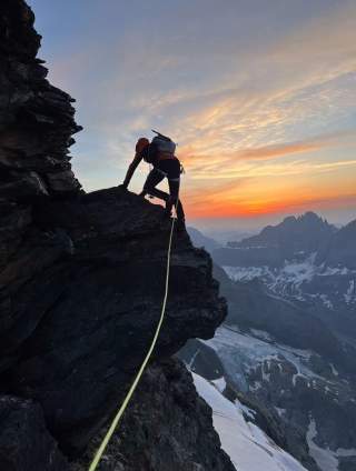 Bergsteiger klettert an einer Felswand, gesichert mit einem Seil, im Hintergrund Sonnenuntergang und schneebedeckte Berge.