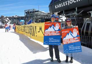 Menschen stehen im Schnee vor einer Seilbahnstation und halten Protestschilder sowie ein großes gelbes Banner mit der Aufschrift „Finger weg von den Gletschern!“.