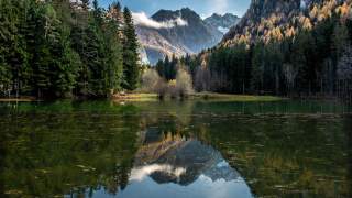 Ein grüner See mit klarem Wasser wird von Nadelbäumen am Ufer geziert. In der Mitte des Bildes ergibt sich der Blick auf die steinernen Gipfel.