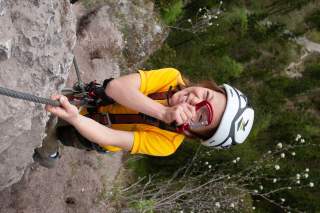 Kind mit Kletterausrüstung und Helm klettert an einem Felsen und hält sich an einem Sicherungsseil fest.