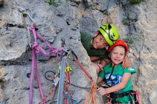 Zwei Kinder mit Kletterhelmen und Gurten umarmen sich an einer Felswand, neben ihnen hängen Karabiner und bunte Kletterseile.