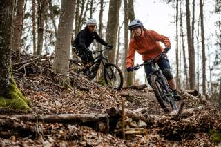Zwei Personen fahren mit Mountainbikes durch einen herbstlichen Wald über einen unebenen, mit Ästen und Laub bedeckten Trail.