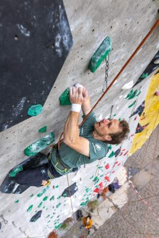 Ein Mann klettert an einer steilen Indoor-Kletterwand mit grünen Griffen, gesichert durch ein Kletterseil.