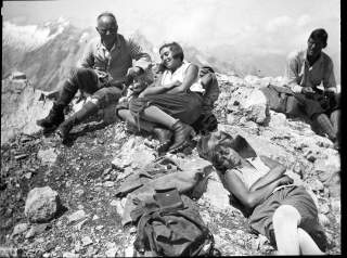 Vier Menschen, davon zwei Frauen und zwei Männer, machen Rast auf dem Gipfel eines Berges. Das Foto ist in schwarz-weiß.