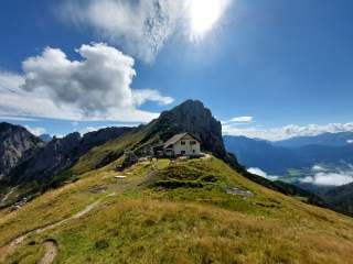 Berghütte auf einer sonnigen Almwiese mit umliegenden Bergen und weiter Aussicht ins Tal.