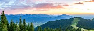 Im Hintergrund sind ganz viele Gipfel zu sehen, die blau vor dem Sonnenuntergangshimmel aussehen. Das Foto zeigt den Blick auf eine grüne Bergkuppe mit viel Wald, auf der breite Wege zu einer Alm führen.