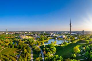 Blick von oben auf den grünen Olympiapark mit Olympiastadion, Olympiasee und Olympiaturm in München.