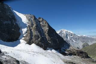 Schneebedeckte Felswand und steile Bergspitzen unter klarem, blauem Himmel. Im Hintergrund ist ein großer, schneebedeckter Berg zu sehen.