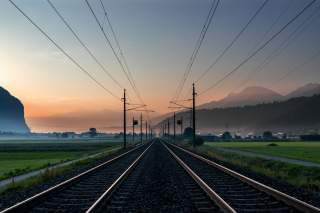 Blick auf Bahngleise, die sich in der Ferne im Licht eines Sonnenaufgangs zwischen grünen Feldern und Bergen verlieren.