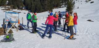 Eine Gruppe von Menschen mit Schneeschuhen steht auf einer verschneiten Wiese vor einer Informationstafel, umgeben von Bäumen und Bergen.